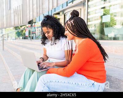 Cheerful diverse female friends in trendy clothes sitting on stair and browsing laptop on city street in summer day Stock Photo