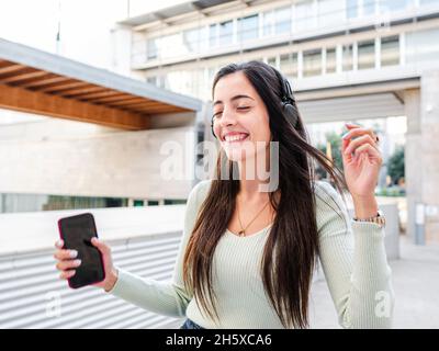 Cheerful young Woman with long brown hair in casual clothes walking with closed eyes and listening to music in headphones on city street in daylight Stock Photo