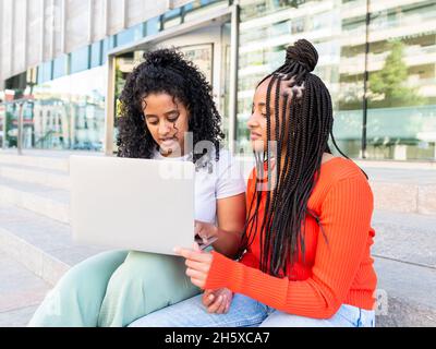 Cheerful diverse female friends in trendy clothes sitting on stair and browsing laptop on city street in summer day Stock Photo