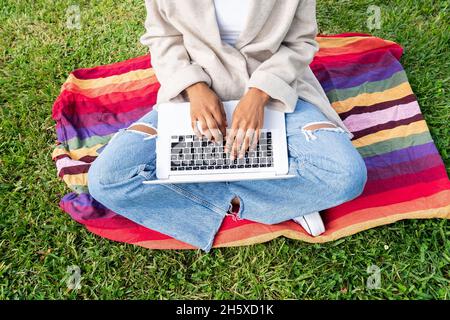 Anonymous African American female typing on modern netbook while sitting on grassy lawn in park Stock Photo