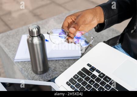 Crop anonymous person with stylish eyeglasses in hand sitting with netbook near thermos of hot drink and copybook on street Stock Photo