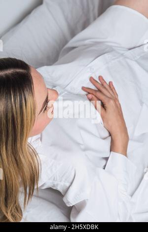 From above young gentle pregnant female touching tummy while lying down on bed Stock Photo