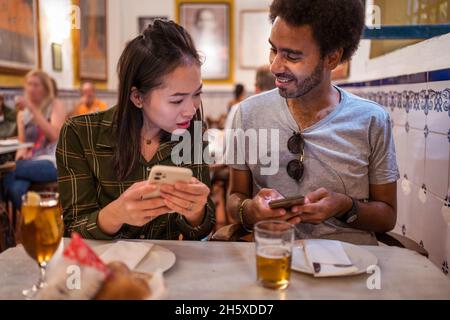 Happy young multiracial couple in casual clothes using smartphones while sitting together at table with glasses of beer in modern restaurant Stock Photo