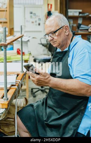 Concentrated senior male master in apron and eyeglasses using mobile phone while sitting at workbench during printing process in studio Stock Photo