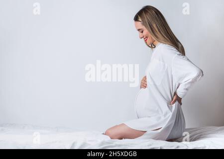 Side view of young gentle female touching tummy while sitting on bed and smiling happily Stock Photo
