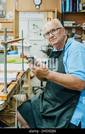 Concentrated senior male master in apron and eyeglasses using mobile phone while sitting at workbench during printing process in studio Stock Photo
