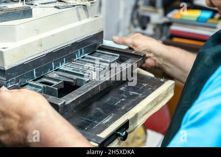 Crop unrecognizable elderly male artisan in apron and eyeglasses preparing metal frame for letterpress printing in studio Stock Photo