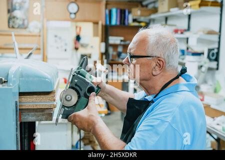 Side view of concentrated senior male carpenter with white hair in apron and eyeglasses cutting wooden boards with circular saw during work in joinery Stock Photo