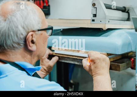 Crop elderly male worker in casual clothes and eyeglasses making marks on pile of paper sheets while operating printing press machine Stock Photo