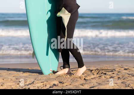Side view of cropped unrecognizable female surfer in wetsuit with surfboard standing looking away on seashore washed by waving sea Stock Photo