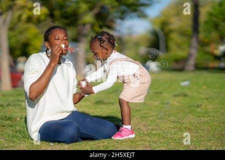 Calm African American Woman sitting on green grass and blowing soap bubbles while playing with daughter in park in summer time Stock Photo