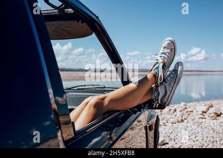 Crop unrecognizable female chilling on passenger seat in car with legs in opened window on stony shore of pond Stock Photo