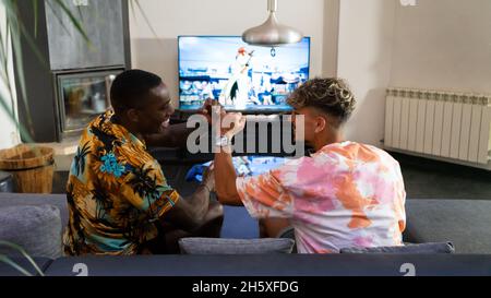 Side view of positive multiethnic male friends doing fist bump and looking at each other while playing video game in living room Stock Photo