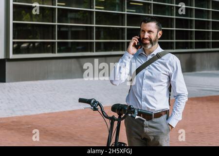 Male worker in formal wear standing with bicycle and having conversation on mobile phone in downtown Stock Photo
