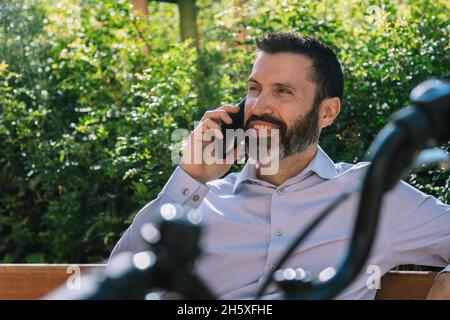 Male worker in formal wear sitting in bench in a park with bicycle and having conversation on mobile phone Stock Photo