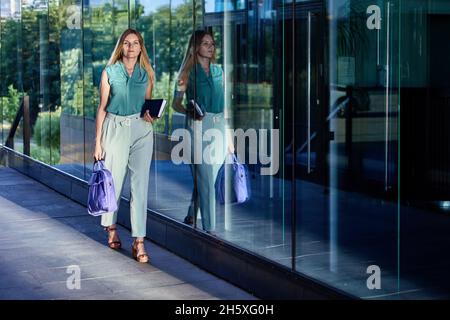 Woman in business suit carries laptop in case and notebook when she walks near glass wall of an office building. Stock Photo