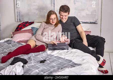 Young couple searches internet using laptop while sitting on bed in bedroom. Stock Photo