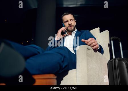 Confident successful and perplexed young European businessman talking on a mobile phone while sitting in an armchair of a hotel lounge, relaxing after Stock Photo