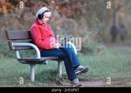 Elderly Senior Caucasian Woman alone sitting on Park Bench wearing Headphones, holding Tablet Stock Photo