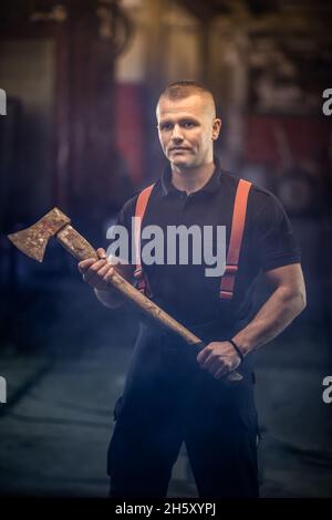 firefighter portrait wearing shirt and red throuser suspenders, holding an axe. smoke and fire trucks in the background. Stock Photo