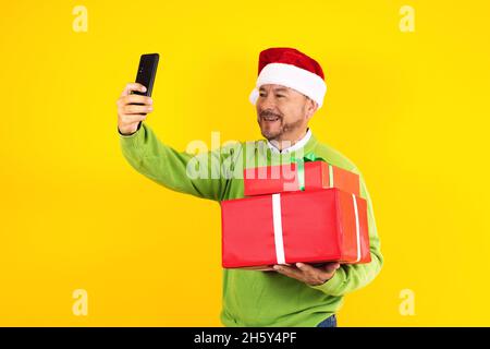 Portrait of senior latin man holding mobile phone taking a photo selfie in a christmas concept on yellow background Stock Photo