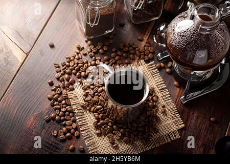 Metallic cup and vacuum coffee maker also known as vac pot, siphon or syphon coffee maker and toasted coffee beans on rustic wooden table Stock Photo