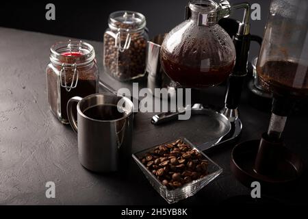 Vacuum coffee maker also known as vac pot, siphon or syphon coffee maker. Metallic cup and toasted coffee beans on rustic black stone table. Stock Photo