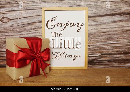 Blackboard with the text ENJOY THE LITTLE THINGS with gift box and red ribbon on wooden background Stock Photo