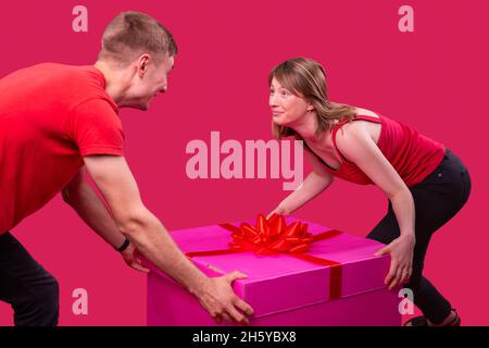 Lovely two persons man and woman wearing red t-shirts and black jeans both holding the big pink present box standing opposite and looking at each Stock Photo
