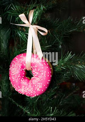 A glazed pink donut hangs from a satin ribbon on a Christmas tree. Sweet Christmas toys. Stock Photo