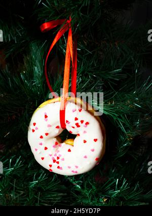 A glazed white donut hangs on a red satin ribbon on a Christmas tree. Sweet Christmas toys. Stock Photo