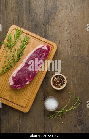 Raw striploin with thym, rosemary, salt and pepper on wooden cutting board. Uncooked gourmet bovine steak. Fresh strip dish short loin. Vertical, top Stock Photo