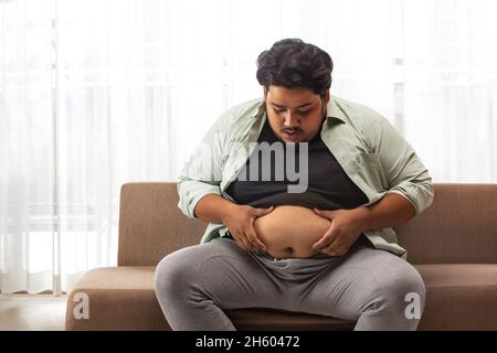 A fat man sitting on couch  holding and looking at extra belly fat. Stock Photo