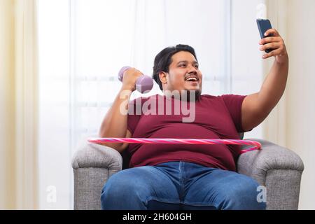 A fat man sitting on couch with dumbles and hulla hoop around his waist clicking selfie. Stock Photo
