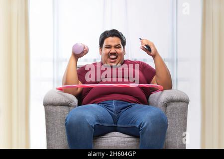 A fat man sitting on couch with dumbles and mobile with a hulla hoop around his waist. Stock Photo