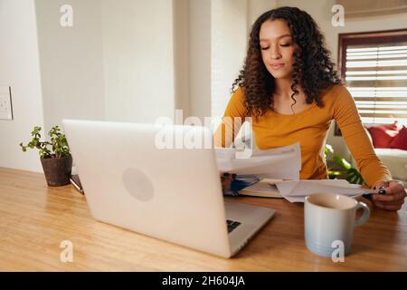 Multi-cultural female paying bills from home. Young professional remote working with laptop in modern apartment Stock Photo