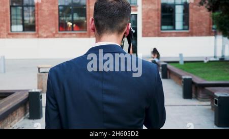 Back front of businessman going to startup office building talking at smarphone discussing company problem with remote collegue explaining business strategy. Outdoor concept Stock Photo