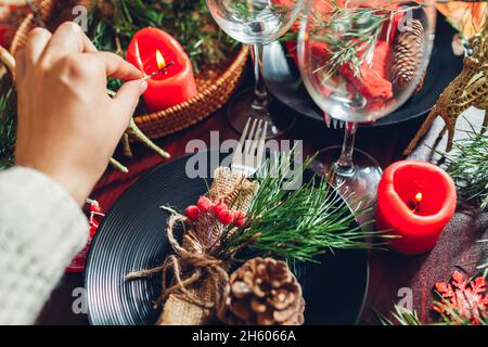 Festive Christmas dinner table setting for two decorated with fir branches, red candles, cones, ornaments. Woman lights candle with burning match Stock Photo