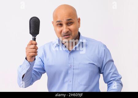 Portrait of a bald man with shaved head holding comb with frustration against white background. Stock Photo