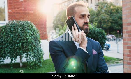 Portrait of leader man in diplomatic suit talking at phone discussing financial strategy with remote collegue. Executive manager standing outside in front of startup building office Stock Photo