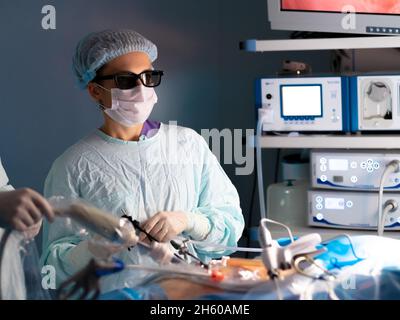 A female surgeon wearing 3D glasses operates on a patient. Laparoscopic minimally invasive surgery. Doctor in surgical uniform, mask and 3D glasses. Modern medical equipment on a blurred background. Stock Photo