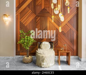 Modern white feather armless chair, squared coffee table, and short wooden planter with green bushes, in a hall with decorated wood cladding wall, and white marble floor Stock Photo