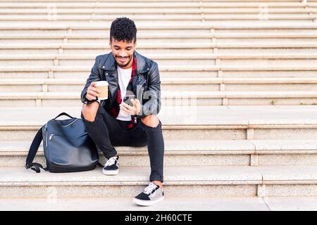 young latin man sitting on a staircase smiling and consulting his phone with a coffee in his hand, concept of technology and urban lifestyle Stock Photo