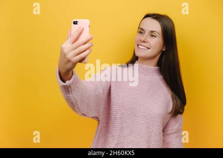 Portrait of nice attractive lovely cheerful funny young woman 20s holding smartphone making selfie, wears pink knitted sweater, isolated over wall Stock Photo