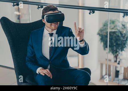 Amazed businessman in formal suit sitting in company lobby wearing virtual reality headset goggles Stock Photo