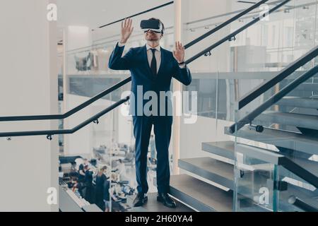Businessman in VR headset stands on staircase in office lobby with hands up in air Stock Photo