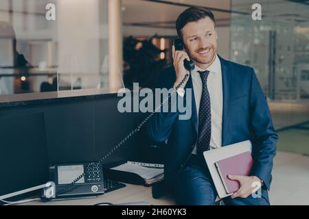 Smiling businessman talking on phone sitting in office on top of desk, holding laptop and agenda Stock Photo