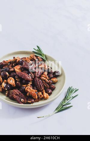 Small plate of roasted honey and rosemary mixed nuts (hazelnut, pecan, cashew) on a white marble backdrop. Stock Photo