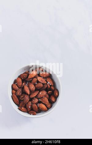 Small bowl filled with roasted smoked paprika and salt almonds on a white marble backdrop. Stock Photo