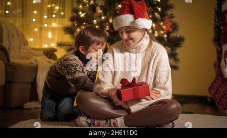 Cheerful smiling boy with mother giving Christmas presents and hugging under Christmas tree at house. Families and children celebrating winter